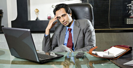Thisimage shoiws a man sitting in his office, in front of his laptopwith is hand on his head. He looks worried and stressed